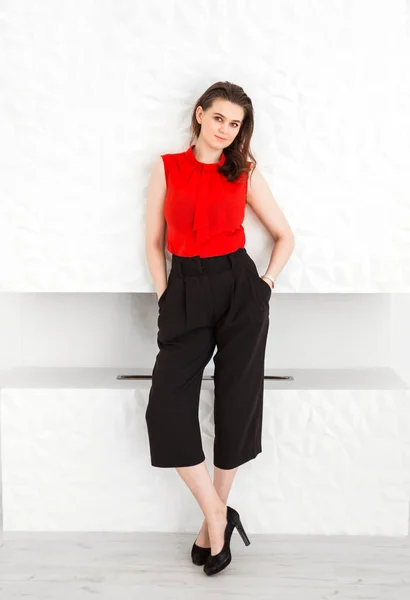 Brunette girl in a red blouse, black capri pants and high-heeled shoes posing against the background of a white fireplace. Vertical photo