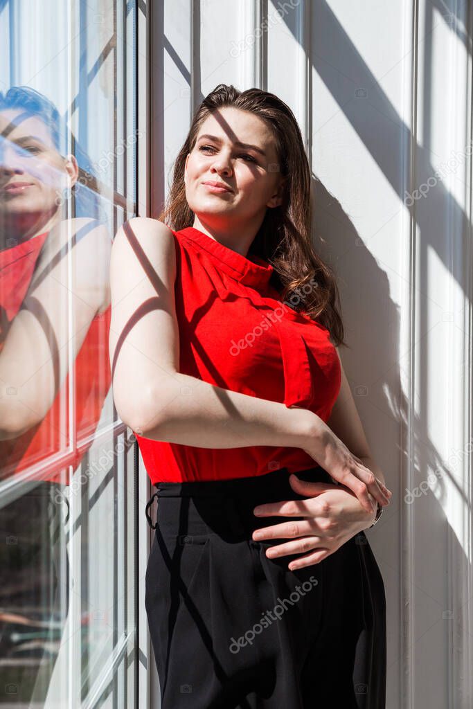 Fashionable brunette girl in a red blouse poses near the window and enjoys the sunlight and warmth. Vertical photo