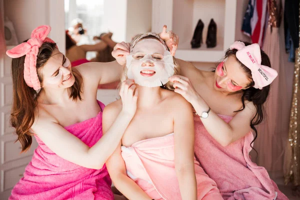 Başlarında kozmetik bandajlarla pembe havlular içinde üç güzel genç kadın kameranın önünde gözlerinin altında bantlarla ve bir peçete kozmetik maskesiyle poz veriyorlar. Kızlar arkadaşlarının kumaş maskesini dikkatlice çıkarıyorlar. Yatay fotoğraf