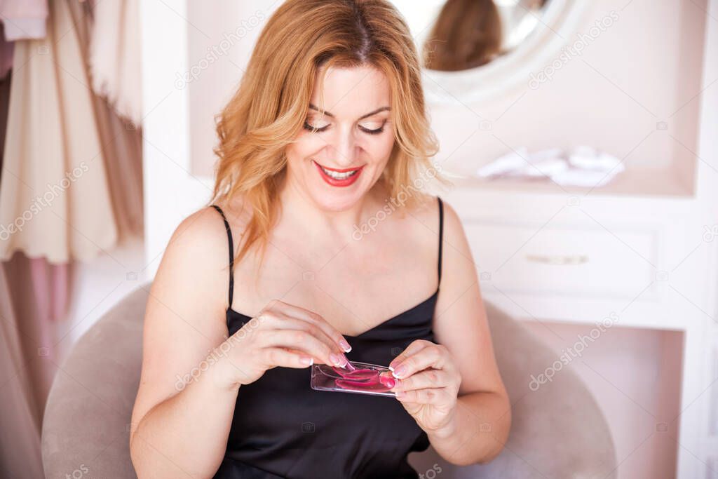 A beautiful young woman takes out pink patches from the package. The woman looks at the patches and smiles. Home care concept. Horizontal photo