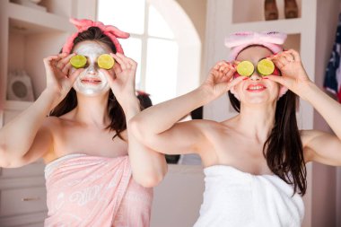 Güzel, mutlu, pozitif kadınlar havluya sarılmış, kafalarında kozmetik bandaj ve gözlerinde limonla. Kızlar gülümser ve kameraya bakar. Yatay fotoğraf