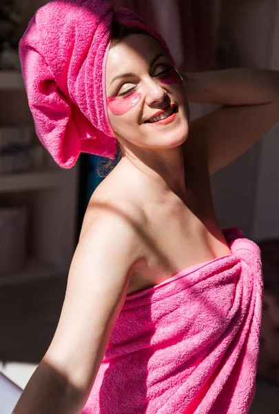 A beautiful and happy woman with a body and hair wrapped in a pink towel and with pink patches under her eyes is posing in a sunny bathroom. Woman smiling with closed eyes. Vertical photo