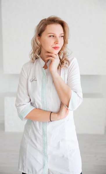 Beautiful young woman doctor in white uniform posing against white wall background and looking at camera. Vertical photo