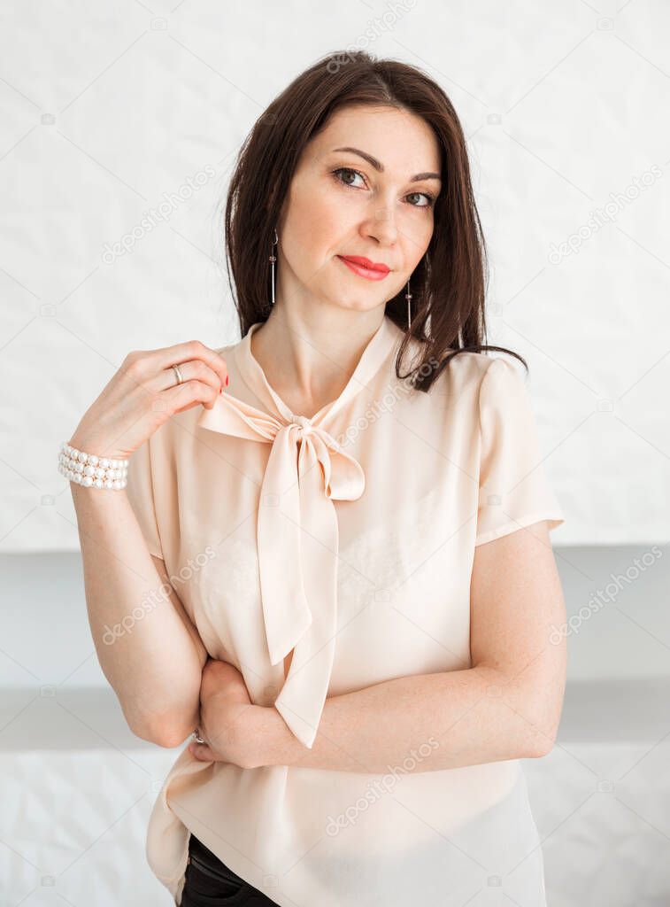 Fashionable brunette girl in a light blouse stands against a white wall, posing and looking at the camera. Vertical photo