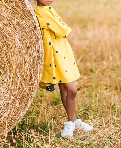 流行の黄色のドレスに身を包んだアフリカ系アメリカ人の少女で 水玉と白い運動用の靴が干し草を背景にポーズをとっている 垂直写真 — ストック写真