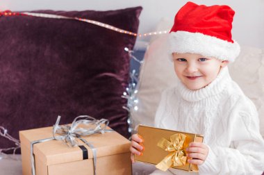 Noel Baba şapkalı küçük çocuk elinde bir hediyeyle kanepede oturuyor. Yatay fotoğraf