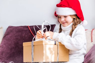 Noel Baba şapkası takan bir kız kanepeye oturur ve içinde yeni yıl hediyesi olan büyük bir kutuyu açar. Kutuya bakan kız. Yatay fotoğraf