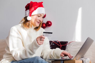 Beyaz kazaklı, Noel Baba şapkalı genç bir kadın bir defter ve kredi kartı kullanarak online alışveriş yapıyor. Yeni yıl alışverişi ve Noel indirimi konsepti. Yatay fotoğraf
