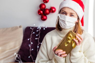 Beyaz kazaklı, Noel Baba şapkalı, tıbbi maskeli ve elinde yeni yıl hediyesiyle genç bir kadın kameraya bakıyor. Yatay fotoğraf