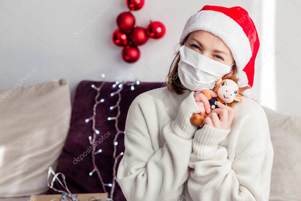 A young woman in a white sweater, Santa Claus hat, medical mask looks into the camera and hugs a soft toy bull. Horizontal photo