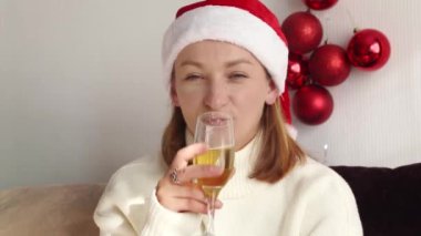 Beyaz kazaklı, kırmızı Noel Baba şapkalı genç bir kadın evinde elinde bir bardak şampanyayla oturuyor kameraya bakıyor ve gülümsüyor. Video