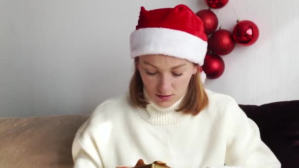 一名身穿白色毛衣 头戴圣诞帽的年轻女子在早上打开了一份新年礼物 — 图库视频影像