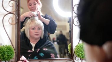 Kısa gri saçlı yaşlı bir kadın güzellik salonunda stil yapıyor. Aynanın önünde oturan yaşlı bir kadın. Video