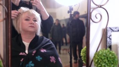 Kısa gri saçlı yaşlı bir kadın güzellik salonunda stil yapıyor. Aynanın önünde oturan yaşlı bir kadın. Video