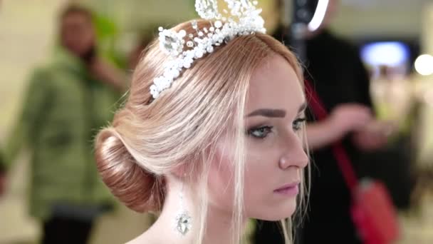 Eine schöne junge Braut mit stylischer Frisur, einem Diadem auf dem Kopf, Ohrringen und einem weißen Kleid sieht sich selbst im Spiegel an — Stockvideo