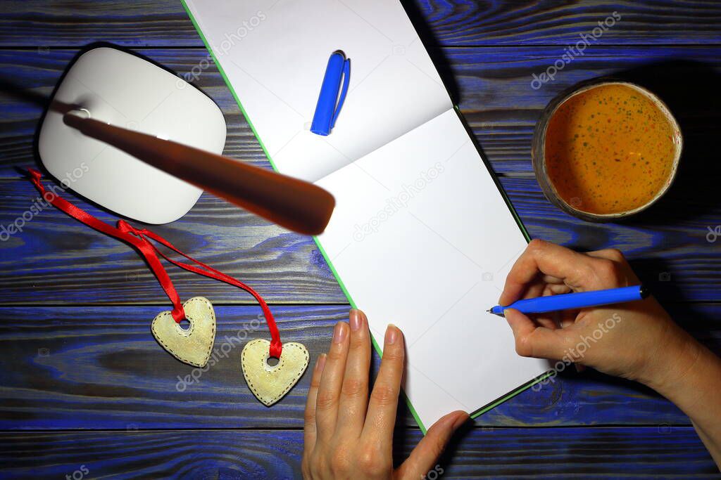 Hands of a girl with pen drawing on blank sheet of open notebook on blue wooden table with white table lamp, pen and cup of tea at night. Top view. ?oncept of creativity and relaxation