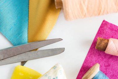 Dikiş, yamalama, terzilik ve moda konsepti - atölyedeki beyaz çalışma masasında yakın çekim araçları, makas, mavi ve pembe iplikler, iğneleme, renkli yama kumaş parçaları, dikey