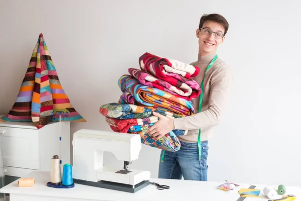 Artesanato, criatividade, conceito de patchwork. pela mesa de trabalho com muitas ferramentas para há um homem bonito jovem com sorriso encantador, ele está segurando amout de colchas coloridas — Fotografia de Stock