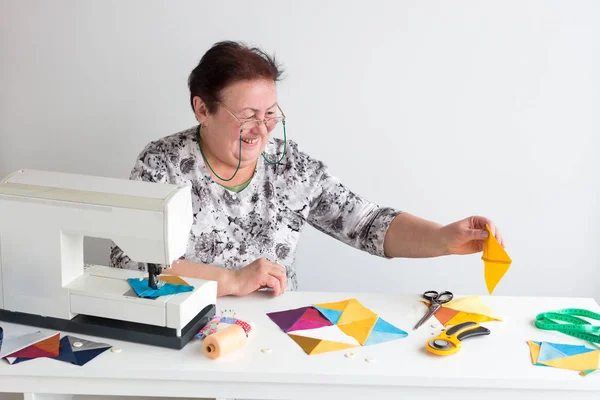 Profesión, fabricación, concepto de acolchado. hay una vieja mujer sonriente en camisa blanca con patrón floral y gafas, ella está sentada en el escritorio con máquina de coser y recoger parches juntos — Foto de Stock