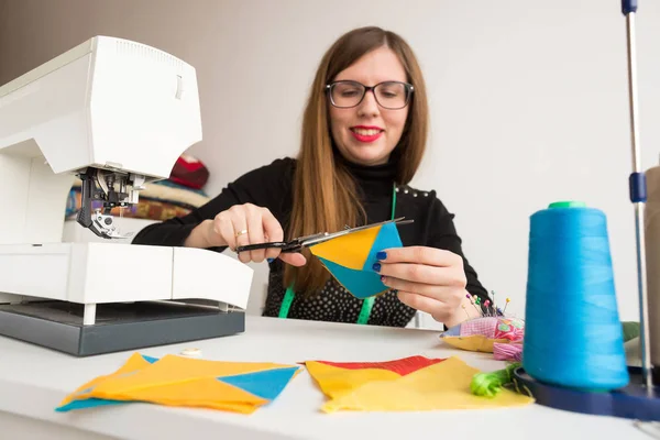 Techniek, naaien, regeling concept. vergadering wordt om een tabel van de jonge vrouw met lange haren en heldere rode lippen werkt crafting quilt gemaakt van verschillende patches in gele en blauwe kleuren — Stockfoto