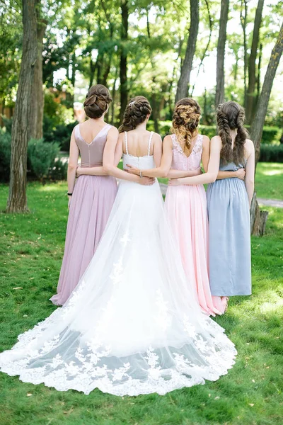 Feest, betrokkenheid, bruidsmeisje concept. ruggen van vier jonge slanke mooie vrouwen, gelukkige bruid in traditiebewust witte jurk en haar bridemaids, ze lopen op het park vol bomen en struiken — Stockfoto