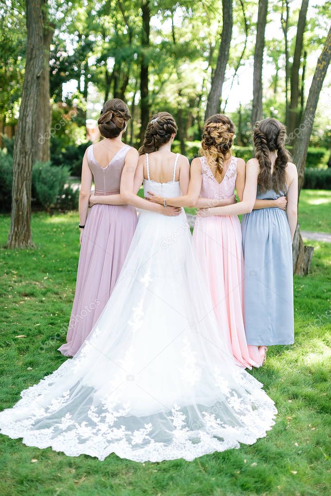 celebration, engagement, bridesmaid concept. backs of four young slim beautiful women, happy bride in traditionaly white dress and her bridemaids, they are walking on the park full of trees and bushes