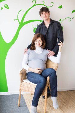 Aile, bağlantı, ilişki kavramı. Duvarın arka planında yeşil ağaç resmi olan genç bir çift ebeveyn olmaya hazırlanıyor, hamile güzel kadın sandalyede oturuyor.