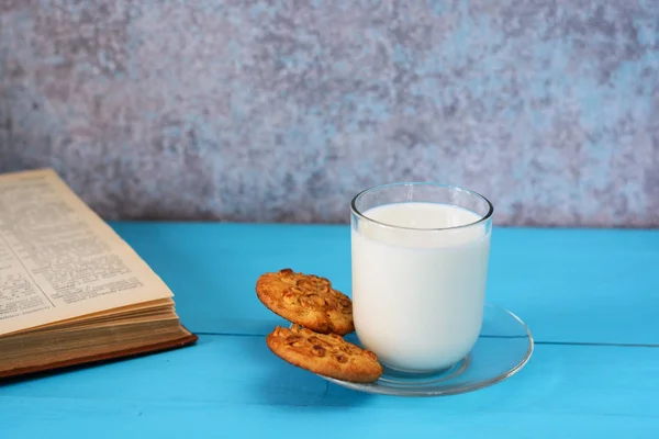 A glass of milk, homemade cakes (oatmeal cookies with nuts) and a book on a blue wooden background.