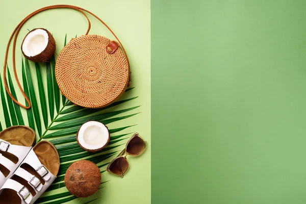 Round rattan bag, coconut, birkenstocks, palm branches, sunglasses on green background. Banner. Top view with copy space. Trendy bamboo bag and shoes. Summer fashion flat lay. Trip, vacation concept