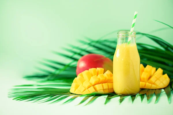 Delicious juicy smoothie with orange fruit and mango on turquoise background. Copy space. Pop art design, creative summer concept. Fresh juice in glass bottles over green palm leaves. — Stock Photo, Image