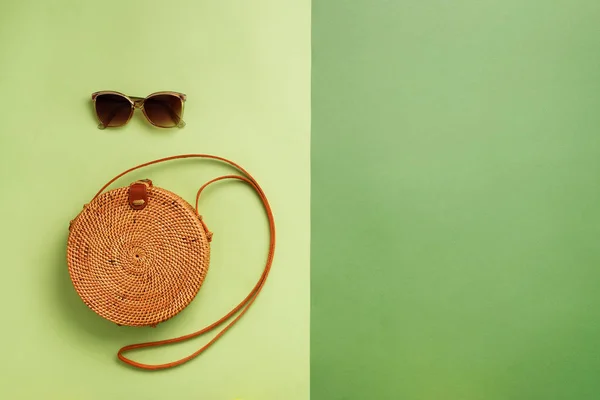 Round rattan bag, sunglasses on green background. Banner. Top view with copy space. Trendy bamboo bag and white shoes. Summer fashion flat lay. Trip, vacation concept