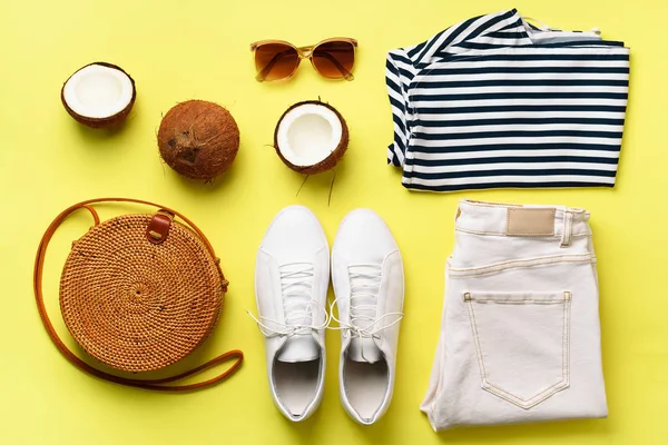 Female white sneakers, jeans, striped t-shirt, rattan bag, coconut and sunglasses on yellow background with copy space. Top view. Summer fashion, capsule wardrobe concept. Creative flat lay.