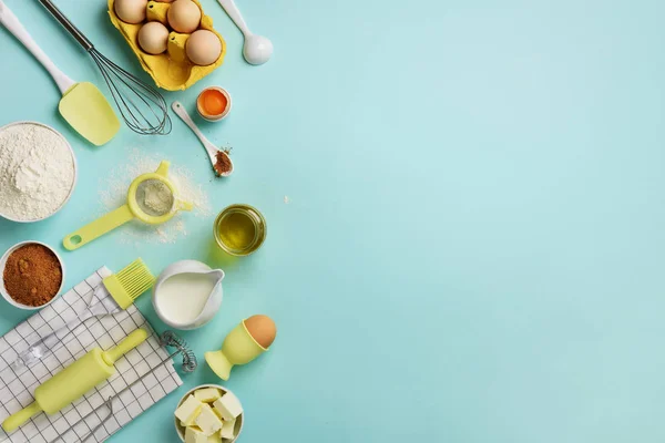 Butter, sugar, flour, eggs, oil, spoon, rolling pin, brush, whisk, towel over blue background. Bakery food frame, cooking concept. Ingredients on kitchen table. Top view, copy space. Flat lay.