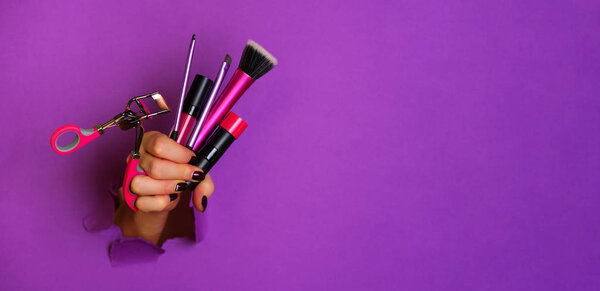 Woman hand with professional cosmetic tools for make up: brushes, mascara, lipstick, eyelash curler on violet background. Beauty concept. Banner for cosmetics sale. Copy space.