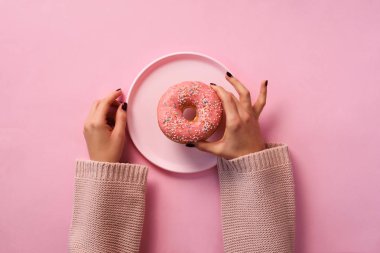 Kadın eli, pembe arka planda donutu tabakta tutuyor. Üst manzara, düz yatış. Tatlı, tatlı, diyet konsepti. Fotokopi alanı olan pankart. Tatilden sonra kilo kaybı