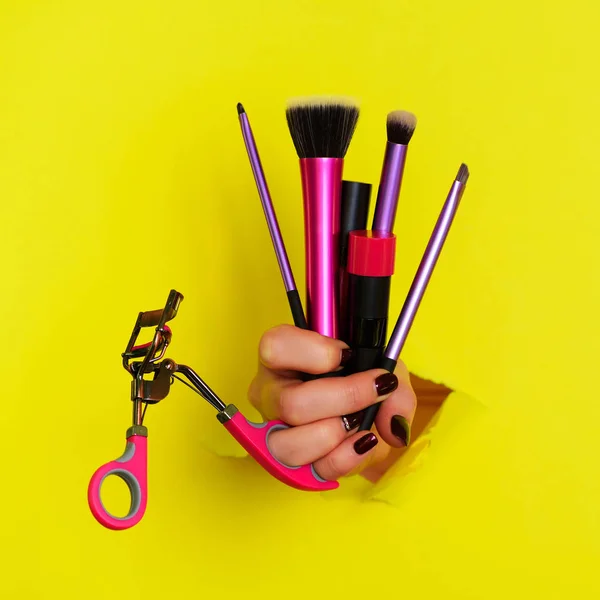 Main de femme avec des outils cosmétiques professionnels pour le maquillage : brosses, mascara, rouge à lèvres, friseur de cils sur fond jaune. Concept de beauté pour la vente de cosmétiques. Culture carrée — Photo