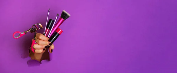 Mano de mujer con herramientas cosméticas profesionales para maquillaje: pinceles, rímel, lápiz labial, rizador de pestañas sobre fondo violeta. Concepto de belleza. Banner para la venta de cosméticos. Copiar espacio —  Fotos de Stock