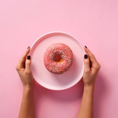 Kadın eli, pembe arka planda donutu tabakta tutuyor. Üst manzara, düz yatış. Tatlı, tatlı, diyet konsepti. Fotokopi alanı olan pankart. Tatilden sonra kilo verdim. Kare mahsul