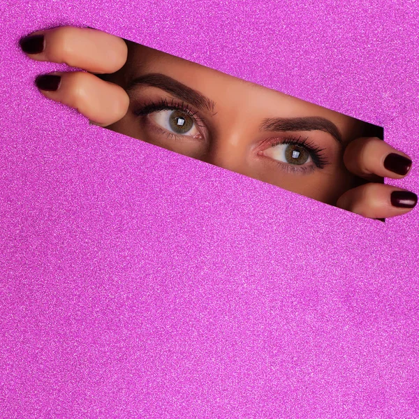 Girl with bright make up looking through hole in violet paper background. Spying, glancing concept. Make up artist, beauty trend. Square crop. Beauty salon advertising banner with copy space
