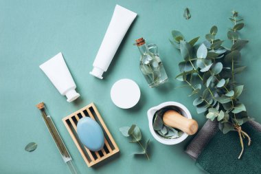 Beyaz kozmetik şişeleri, okaliptüs çiçekleri, havlular, yeşil arka planda sabun. Üst manzara, düz yatış. Doğal organik güzellik ürünü kavramı. Spa, cilt bakımı, vücut bakımı