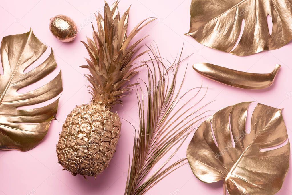 Golden exotic fruits, tropical palm, monstera leaves on pink background. Top view. Flat lay. Food concept. Creative layout of gold pineapple, banana, lemon with copy space.