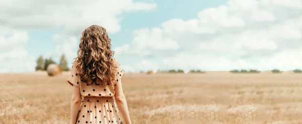 Mulher em vestido de bolinhas bege com cabelo encaracolado, chapéu de palha em pé no campo colhido com fardos de palha. Agricultura fundo com espaço de cópia . — Fotografia de Stock