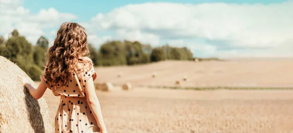 Mulher em vestido de bolinhas bege com cabelo encaracolado, chapéu de palha em pé no campo colhido com fardos de palha. Agricultura fundo com espaço de cópia . — Fotografia de Stock