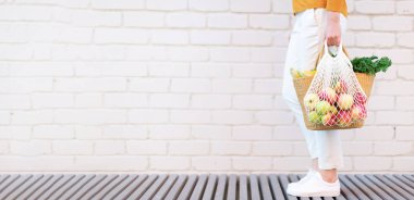 Kopya alanı ile sıfır atık kavramı. Elinde saman sepeti olan ve tekrar kullanılabilen, elmalı, sebzeli, beyaz tuğlalı bir çanta taşıyan bir kadın. Çevre dostu ağ tüccarı. Boşluğu kopyala