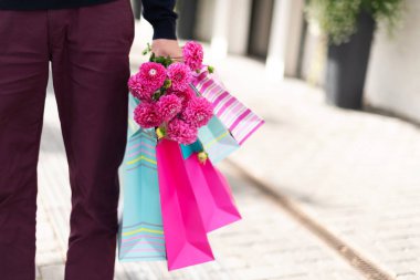 Erkeklerin ellerinde pembe çiçekler ve tuğla arka planda alışveriş torbaları. Doğum günü, Anneler, Kadınlar, Düğün Günü konsepti. Bir deste kağıt hediye çantası.