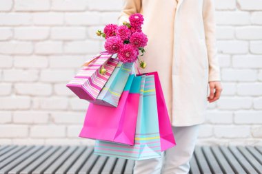 Kadın ellerinde pembe çiçekler ve tuğla arka planda alışveriş torbaları. Doğum günü, Anneler, Kadınlar, Düğün Günü konsepti. Bir deste kağıt hediye çantası.