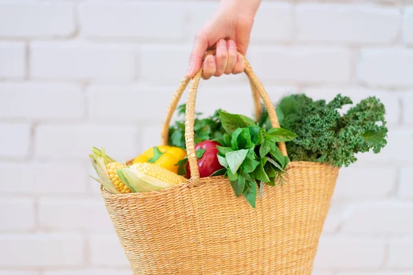 Mujer mano celebración cesta de paja con verduras orgánicas sobre fondo de ladrillo. Comida saludable, dieta vegetariana. Ecológico, cero residuos, concepto libre de plástico . — Foto de Stock