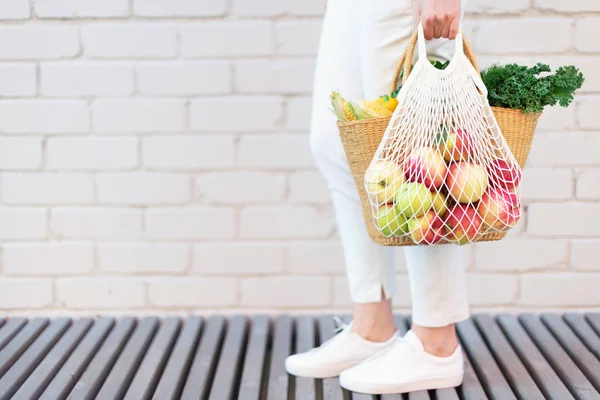 Ragazza che tiene borsa a rete riutilizzabile piena di mele, cesto di paglia con verdure biologiche. Bandiera. Ricevuto. Raccolta autunnale. Stile di vita ecosostenibile. Zero rifiuti, concetto di plastica libera . — Foto Stock