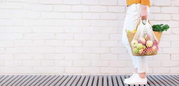 Concepto de cero residuos con espacio de copia. Mujer sosteniendo cesta de paja y bolsa de compras de malla reutilizable conmanzanas, verduras, fondo de ladrillo blanco. Comprador de malla ecológico. Copiar espacio — Foto de Stock