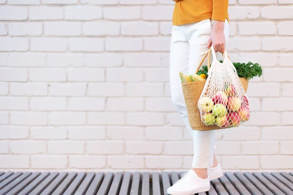 Chica sosteniendo bolsa de compras de malla llena de manzanas y bolsa de paja con verduras orgánicas, fondo de ladrillo. Cero residuos, concepto libre de plástico. Estilo de vida sostenible. Copiar espacio — Foto de Stock